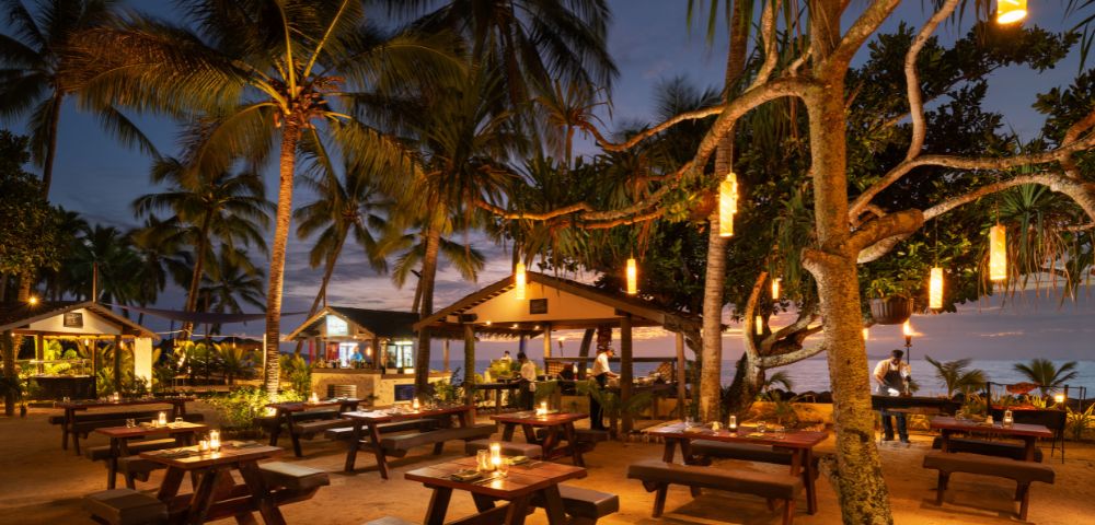 Outdoor beach restaurant at dusk with wooden tables lit by candlelight. Surrounded by palm trees and lanterns, creating a warm, inviting atmosphere.