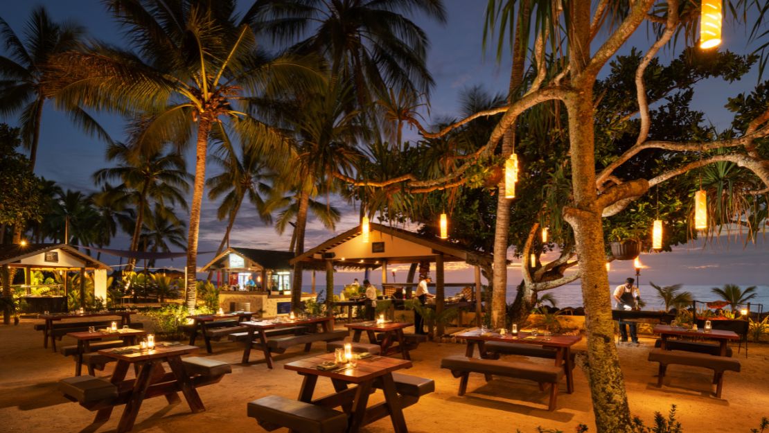 Outdoor beach restaurant at dusk with wooden tables lit by candlelight. Surrounded by palm trees and lanterns, creating a warm, inviting atmosphere.