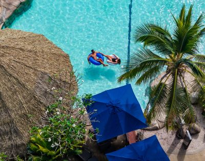 Aerial view of a serene tropical pool with two people on a float. Surrounded by a straw hut, palm tree, and blue umbrellas, evoking relaxation.