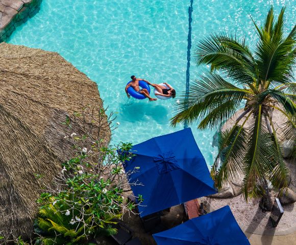 Aerial view of a serene tropical pool with two people on a float. Surrounded by a straw hut, palm tree, and blue umbrellas, evoking relaxation.