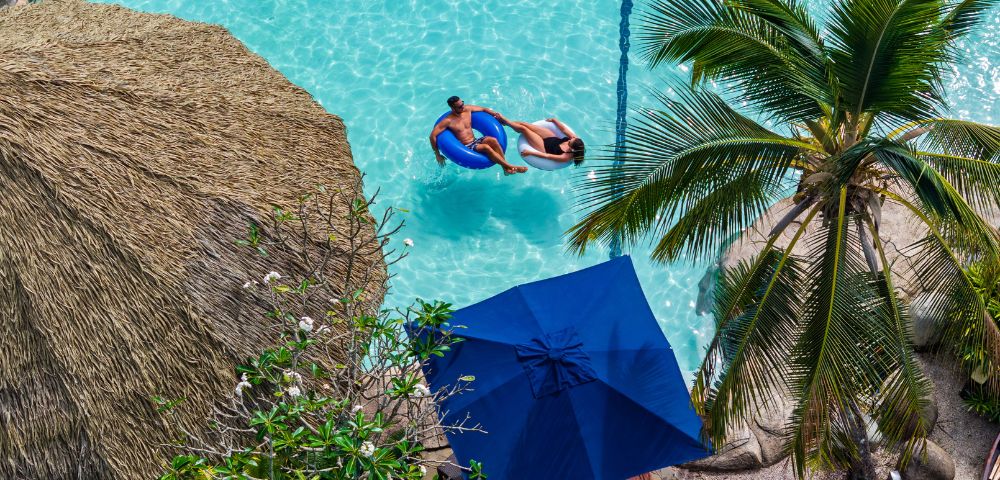 Aerial view of a serene tropical pool with two people on a float. Surrounded by a straw hut, palm tree, and blue umbrellas, evoking relaxation.