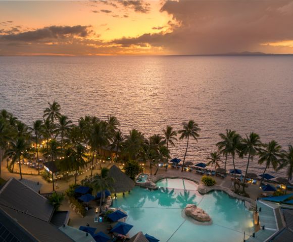 Tropical resort at sunset with palm trees lining a large pool, blue umbrellas, and a warm glow. The ocean and colorful sky create a serene atmosphere.
