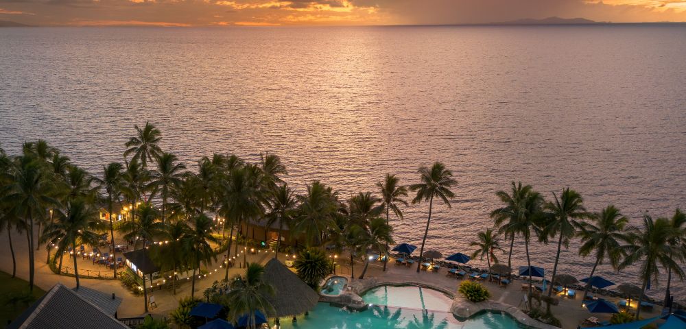 Tropical resort at sunset with palm trees lining a large pool, blue umbrellas, and a warm glow. The ocean and colorful sky create a serene atmosphere.