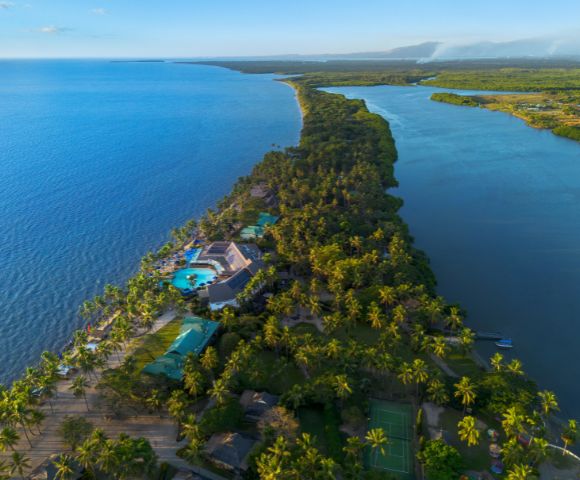 Aerial view of a lush, narrow peninsula with tropical palm trees, a large hotel with blue pools, surrounded by turquoise ocean and calm lagoon waters. Sunny and serene.