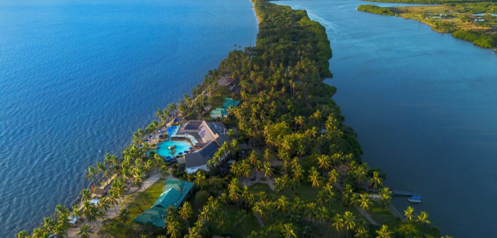 Aerial view of a lush, narrow peninsula with tropical palm trees, a large hotel with blue pools, surrounded by turquoise ocean and calm lagoon waters. Sunny and serene.