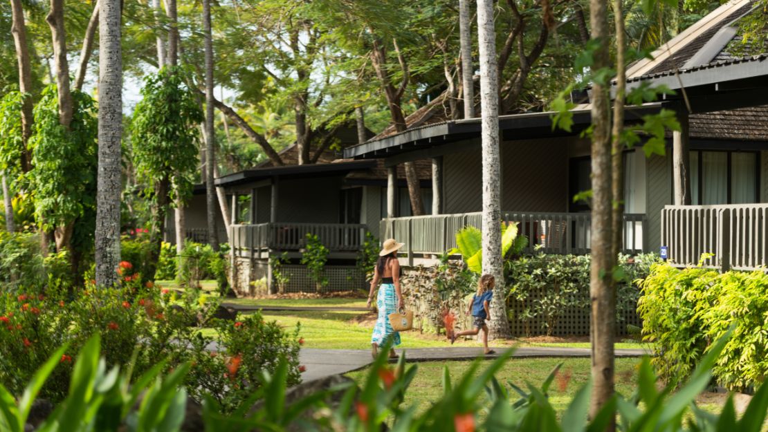 A woman and child walk on a path in a lush, tropical garden. They're near bungalow-style houses, surrounded by green trees and vibrant flowers, conveying tranquility.