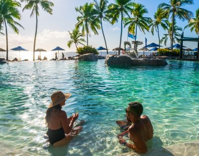 A man and woman relax in a tropical pool, surrounded by palm trees under a sunny sky. Beach chairs and umbrellas line the background, creating a serene, vacation atmosphere.