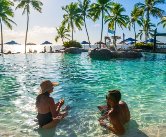 A man and woman relax in a tropical pool, surrounded by palm trees under a sunny sky. Beach chairs and umbrellas line the background, creating a serene, vacation atmosphere.
