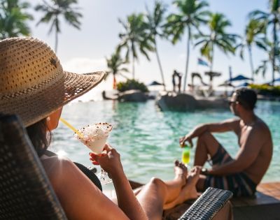 Woman in a sun hat relaxing by a tropical pool with a cocktail, looking at a man sitting nearby. Palm trees and blue sky create a serene, vacation vibe.