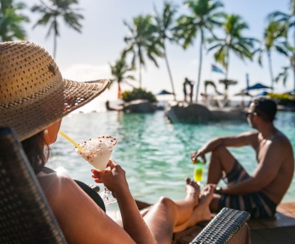 Woman in a sun hat relaxing by a tropical pool with a cocktail, looking at a man sitting nearby. Palm trees and blue sky create a serene, vacation vibe.