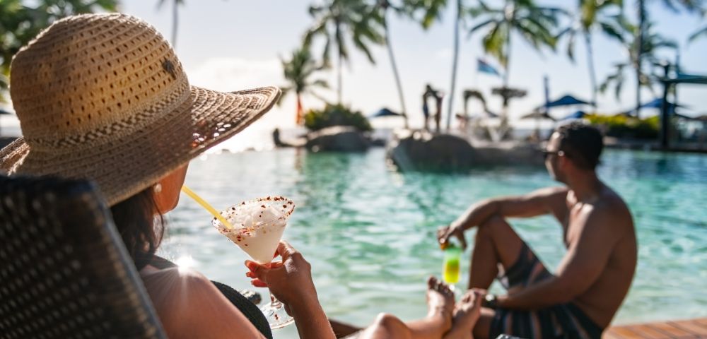 Woman in a sun hat relaxing by a tropical pool with a cocktail, looking at a man sitting nearby. Palm trees and blue sky create a serene, vacation vibe.