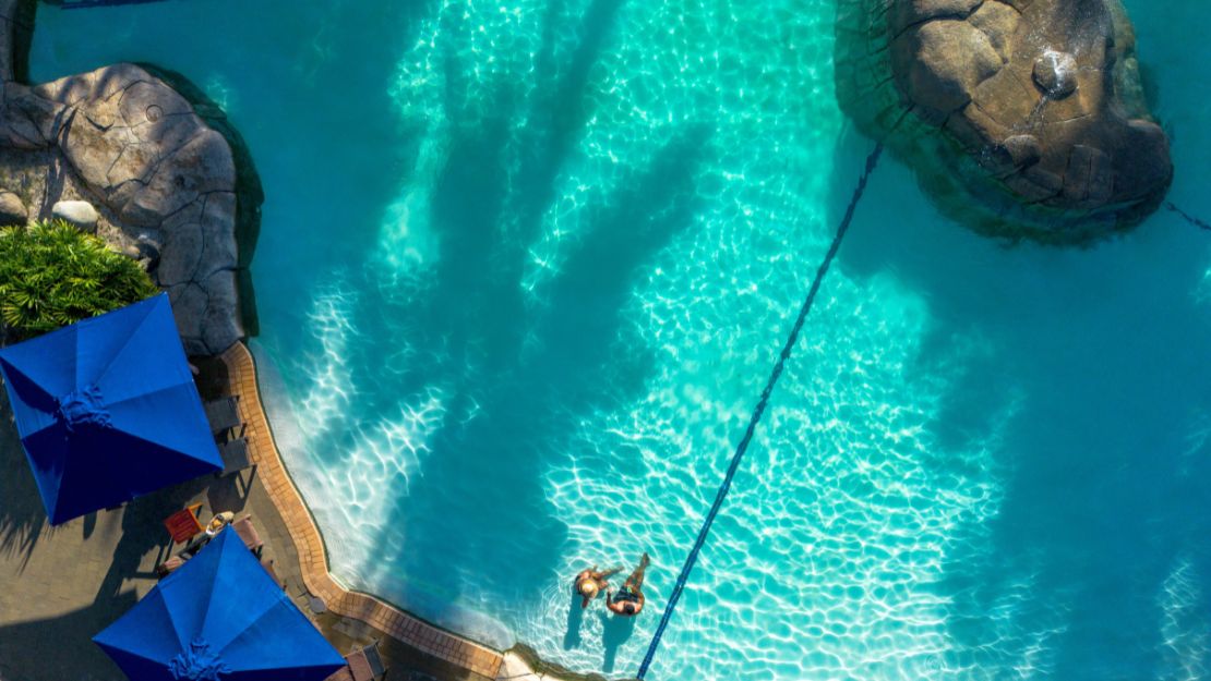 Aerial view of a bright blue swimming pool with rock formations and lounging area. Shadows and blue umbrellas create a serene, vacation-like atmosphere.