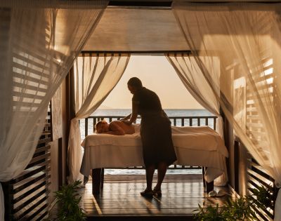 A person receives a relaxing massage on a bed in an open-air cabana with sheer curtains. The serene setting overlooks a calm ocean at sunset.