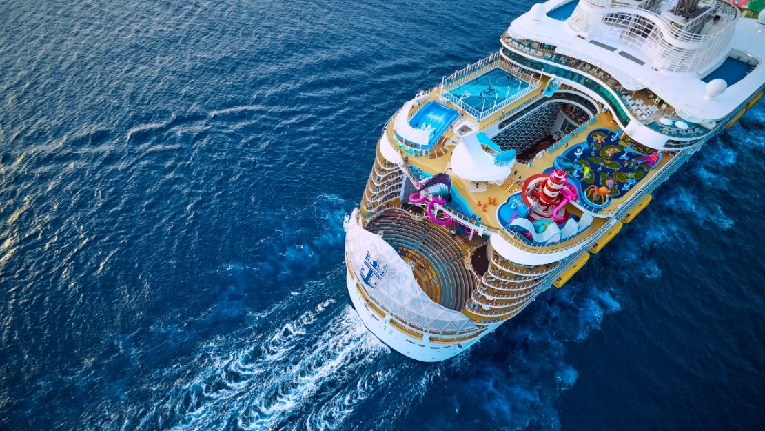 Aerial view of a large cruise ship sailing through deep blue waters, featuring pools, slides, and vibrant deck areas.