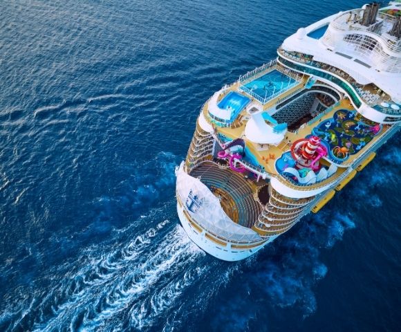 Aerial view of a large cruise ship sailing through deep blue waters, featuring pools, slides, and vibrant deck areas.
