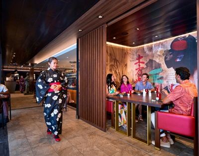 A restaurant scene featuring a woman in a traditional kimono walking by, while a group enjoys their meal at a table.