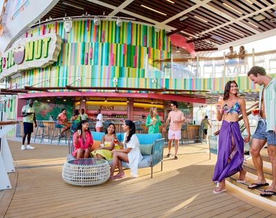 A vibrant outdoor deck on a cruise ship, featuring groups of people socialising near a colorful bar and relaxed seating areas.