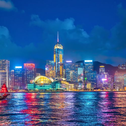 Hong Kong skyline at night with illuminated skyscrapers reflecting on the harbor. A red-sailed boat glides across the vibrant, colorful water.