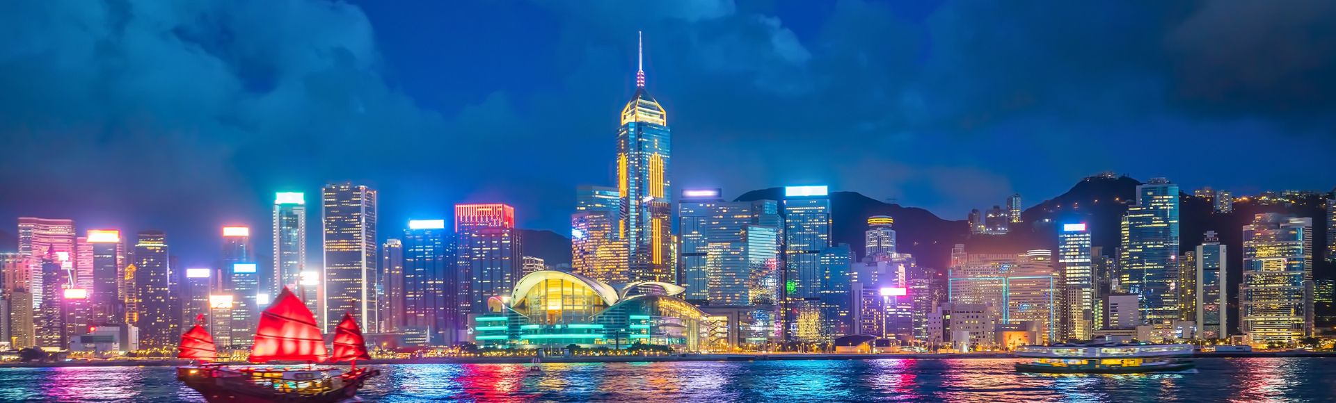 Hong Kong skyline at night with illuminated skyscrapers reflecting on the harbor. A red-sailed boat glides across the vibrant, colorful water.
