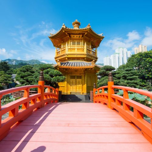 A bright orange arched bridge leads to a golden pagoda surrounded by lush greenery. Skyscrapers rise in the background under a clear blue sky.