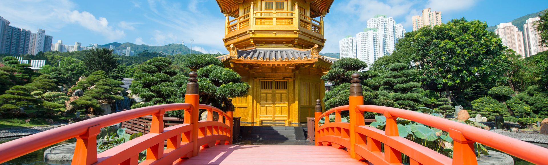 A bright orange arched bridge leads to a golden pagoda surrounded by lush greenery. Skyscrapers rise in the background under a clear blue sky.