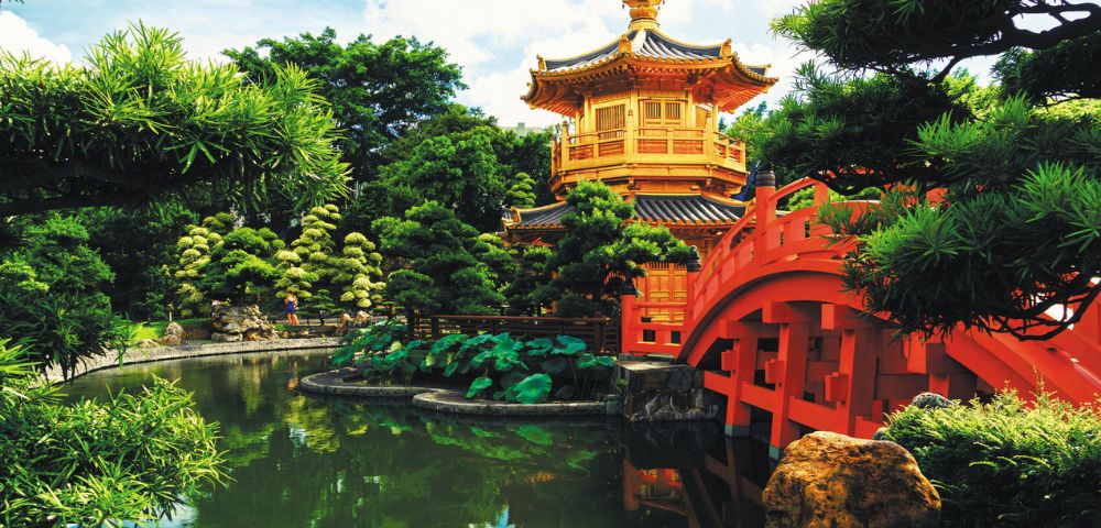 Traditional Chinese pavilion with intricate details, surrounded by lush greenery and a serene pond, featuring a vibrant red arched bridge. Peaceful ambiance.