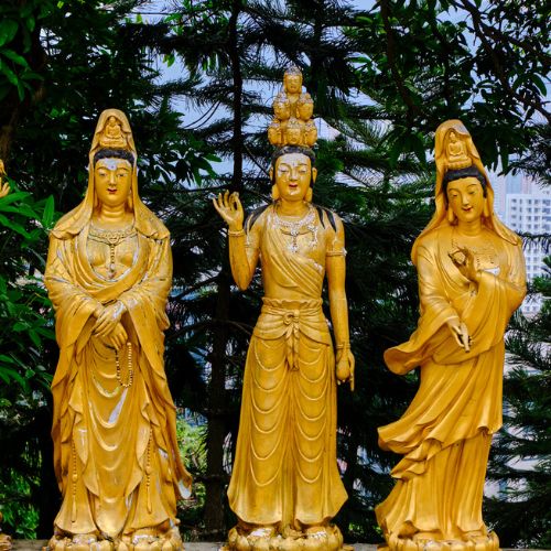 Three golden Buddhist statues stand in serene poses, surrounded by lush greenery. The central statue raises a hand, conveying tranquility and peace.