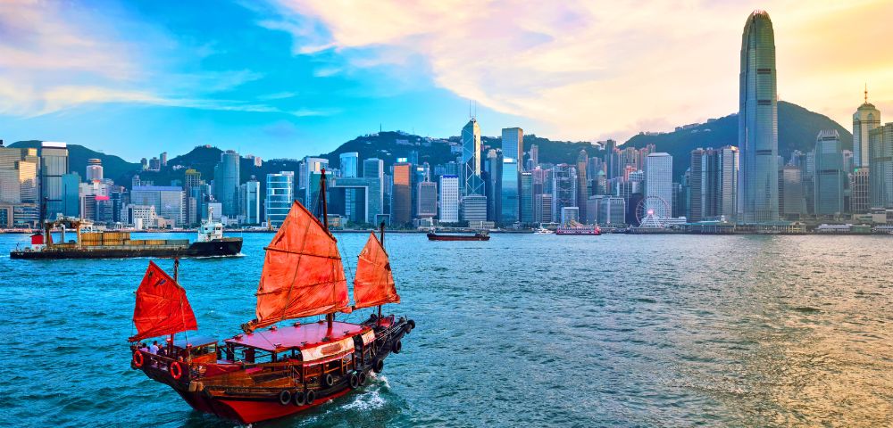 A traditional junk boat with red sails glides across the vibrant blue waters of Victoria Harbour against a backdrop of Hong Kong’s modern skyline at sunset.