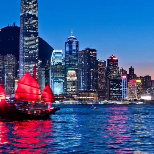 A traditional Chinese junk boat with red sails glides across a shimmering bay at dusk, set against a backdrop of a vibrant, illuminated city skyline.