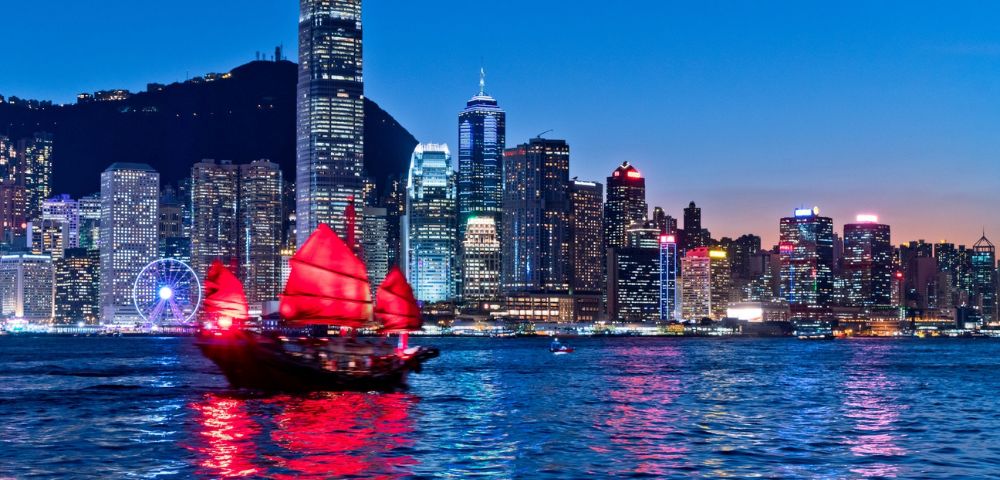A traditional Chinese junk boat with red sails glides across a shimmering bay at dusk, set against a backdrop of a vibrant, illuminated city skyline.