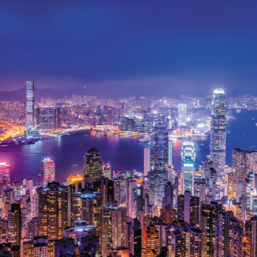 Vibrant nighttime cityscape of Hong Kong with illuminated skyscrapers around Victoria Harbour under a starry sky. The scene conveys an energetic ambiance.