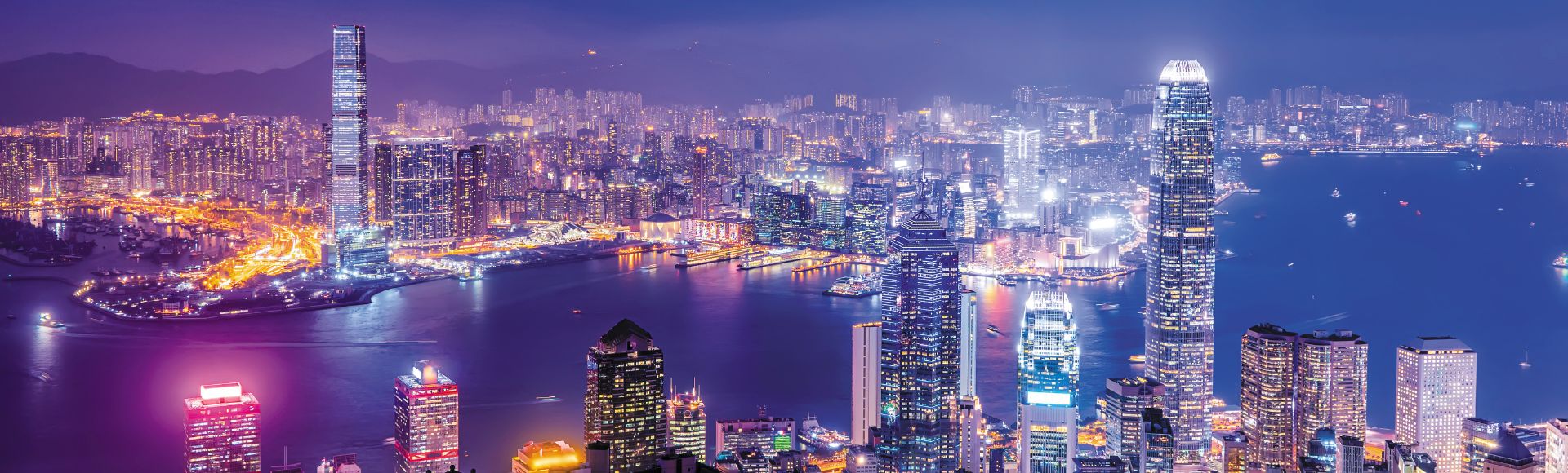 Vibrant nighttime cityscape of Hong Kong with illuminated skyscrapers around Victoria Harbour under a starry sky. The scene conveys an energetic ambiance.