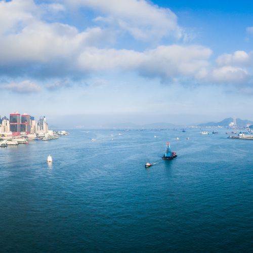 A serene cityscape with a vast blue harbor under a partly cloudy sky. Skyscrapers line the left shore, while boats and ships traverse the expansive waters.