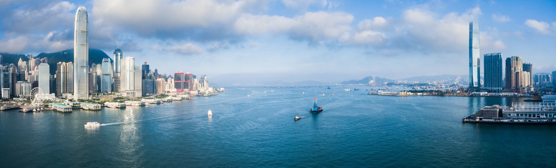 A serene cityscape with a vast blue harbor under a partly cloudy sky. Skyscrapers line the left shore, while boats and ships traverse the expansive waters.