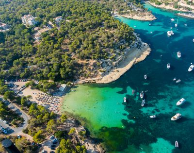 Aerial view of a scenic coastline with turquoise water, anchored boats, a sandy beach, and lush green trees. The atmosphere is serene and inviting.
