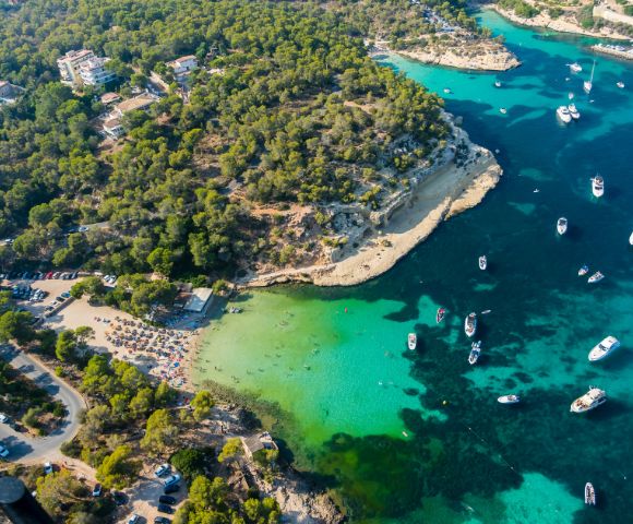 Aerial view of a scenic coastline with turquoise water, anchored boats, a sandy beach, and lush green trees. The atmosphere is serene and inviting.