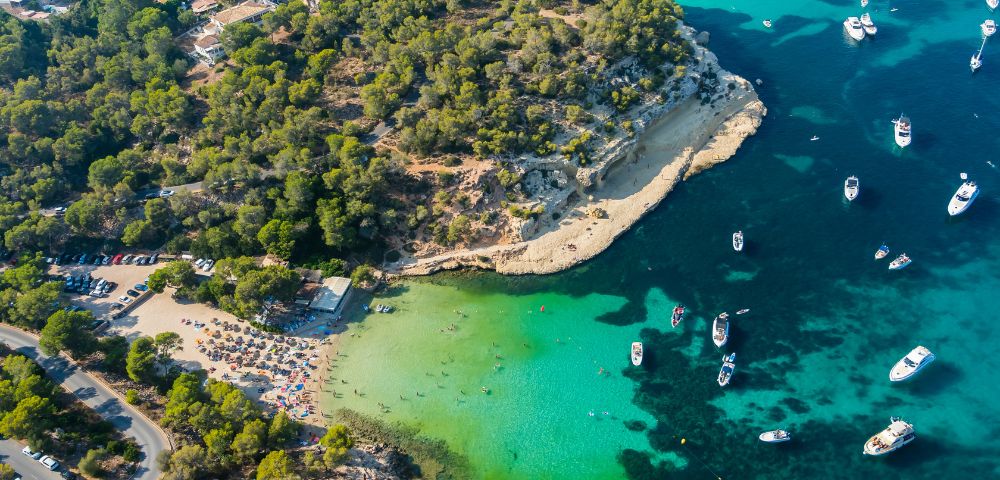 Aerial view of a scenic coastline with turquoise water, anchored boats, a sandy beach, and lush green trees. The atmosphere is serene and inviting.