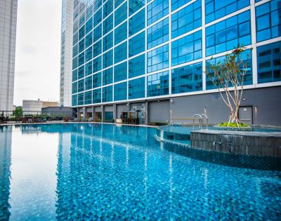 A calm, blue-tiled swimming pool beside a modern glass building. The pool area features a small island with greenery, creating a serene atmosphere.