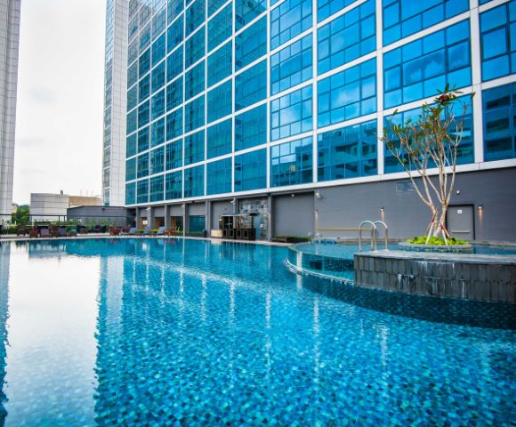 A calm, blue-tiled swimming pool beside a modern glass building. The pool area features a small island with greenery, creating a serene atmosphere.