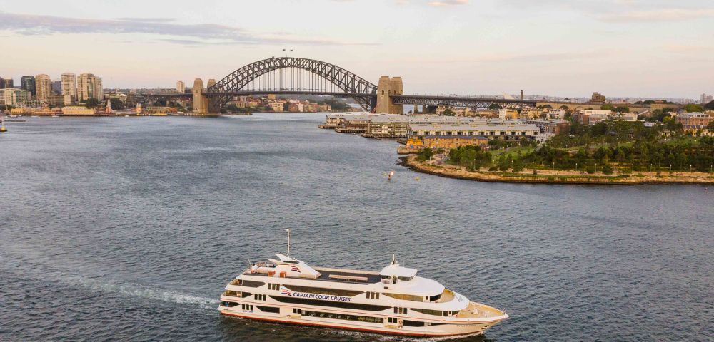 A white cruise ship sails through a wide harbor, passing lush green islands and skyscrapers. In the background, a large steel arch bridge spans two shores.