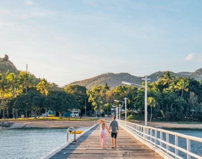 A couple strolls along a sunlit pier, surrounded by lush greenery and distant hills. The scene conveys tranquility and warmth under a clear sky.