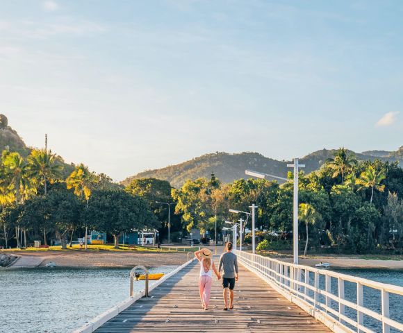 A couple strolls along a sunlit pier, surrounded by lush greenery and distant hills. The scene conveys tranquility and warmth under a clear sky.