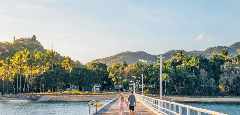 A couple strolls along a sunlit pier, surrounded by lush greenery and distant hills. The scene conveys tranquility and warmth under a clear sky.