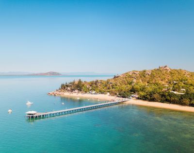 Aerial view of a serene beach with a long pier stretching over turquoise water. Lush green hill on the right, clear sky, and distant islands visible.