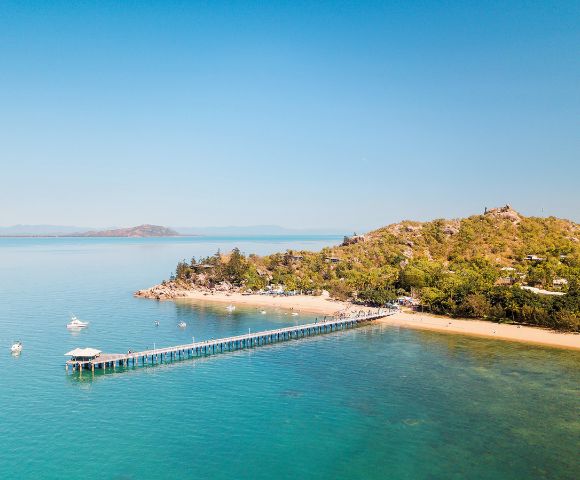 Aerial view of a serene beach with a long pier stretching over turquoise water. Lush green hill on the right, clear sky, and distant islands visible.