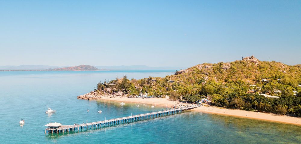 Aerial view of a serene beach with a long pier stretching over turquoise water. Lush green hill on the right, clear sky, and distant islands visible.