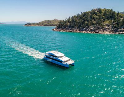 A sleek white and blue yacht glides on clear turquoise waters near a lush, tree-covered island under a bright blue sky, evoking a sense of tranquil adventure.