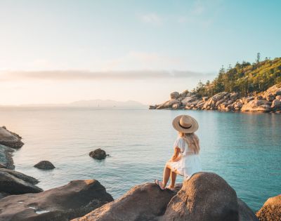 A woman in a white dress and hat sits on rocks overlooking a calm sea. The sky is clear, and a lush, tree-covered coastline is in the background. Tranquil scene.