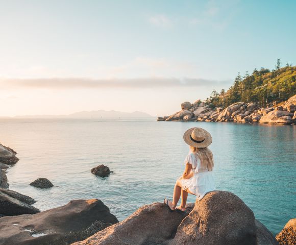 A woman in a white dress and hat sits on rocks overlooking a calm sea. The sky is clear, and a lush, tree-covered coastline is in the background. Tranquil scene.