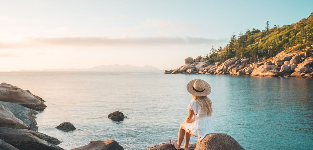 A woman in a white dress and hat sits on rocks overlooking a calm sea. The sky is clear, and a lush, tree-covered coastline is in the background. Tranquil scene.